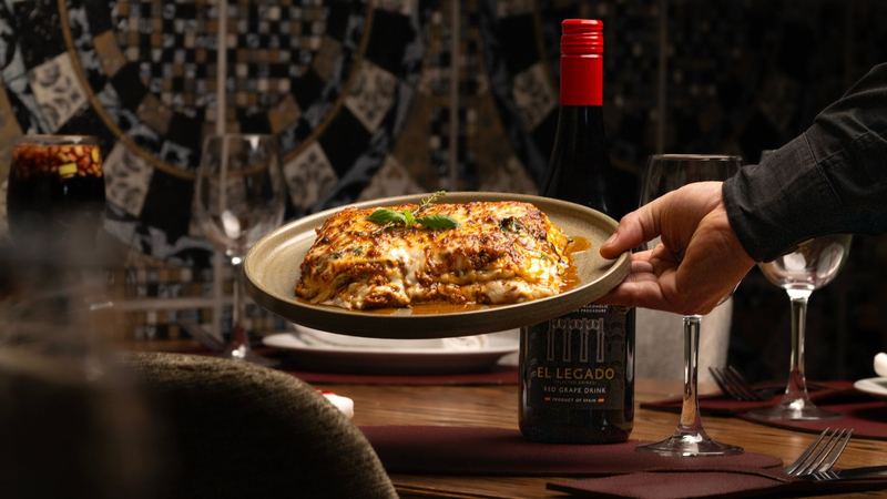 A waiter setting down a plate of baked lasagna beside a bottle and wine glasses