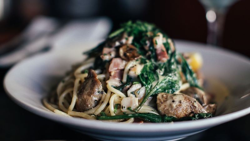 Spaghetti with spinach, mushrooms and pancetta in a white bowl, a wine glass behind