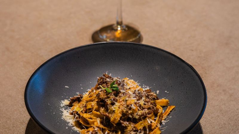 Tagliatelle with a rich ragù and shaved cheese in a dark bowl, a glass beside it