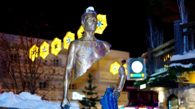 A bronze traveller sculpture in the snow, with holiday lights and boutiques behind