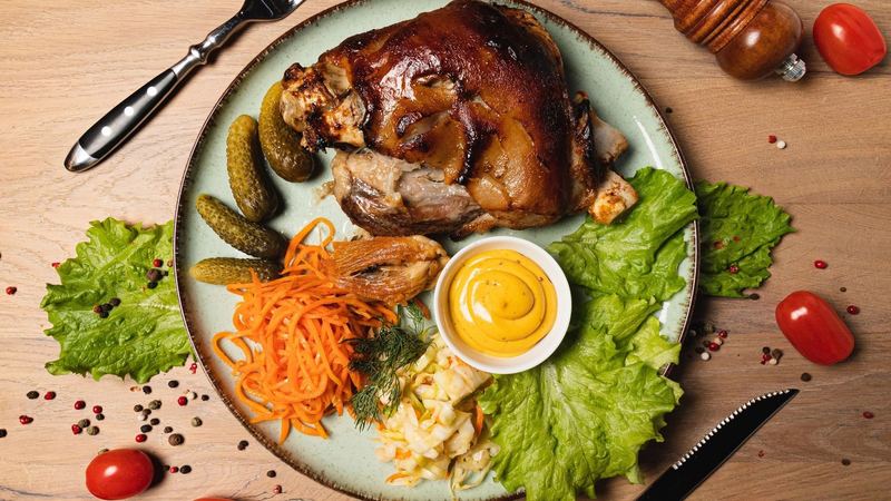 A roast pork knuckle with crackling, mustard, gherkins and cabbage salad on a green plate