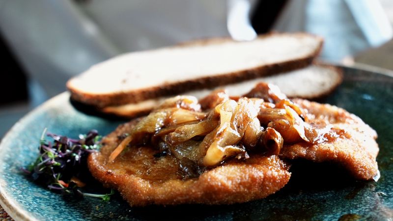 A golden schnitzel topped with caramelised onions, with country bread, on a blue ceramic plate