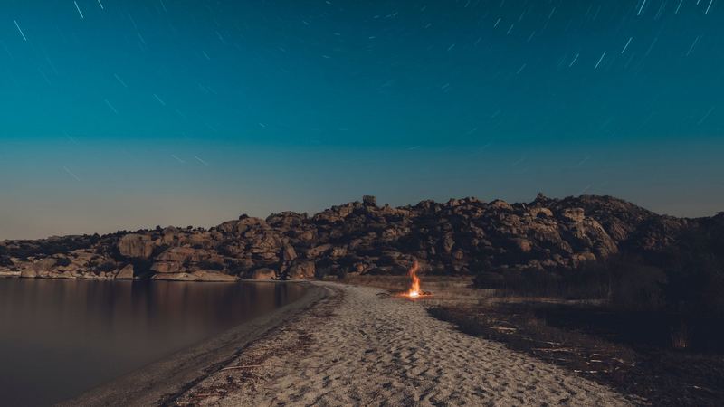 A fire on a sandy spit below granite rocks under a sky of star trails