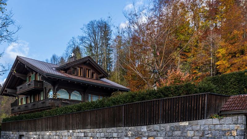 A dark wooden chalet with carved balconies above a stone wall, among autumn trees under a blue sky