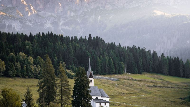 A white village church and a meadow below the pale Dolomite peaks at sunrise