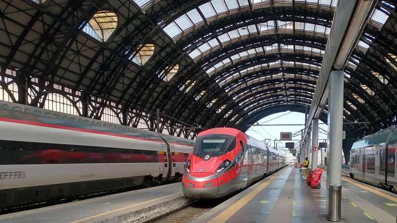 A red Frecciarossa high-speed train under the iron and glass roof of Milano Centrale