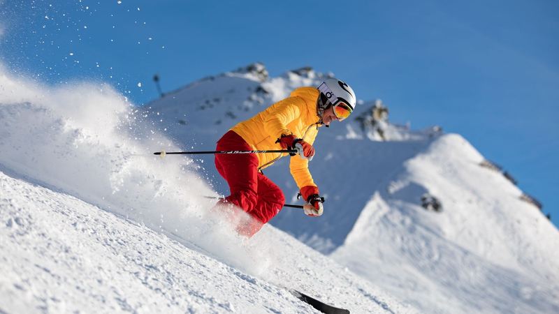 A skier in a yellow jacket and red trousers carving through fresh snow below a white summit