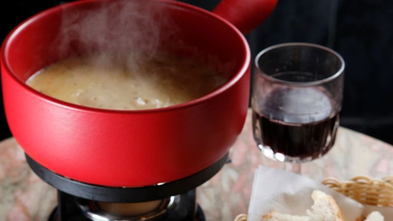 A red pot of cheese fondue steaming on its burner, with bread, red wine and pickles on a marble table