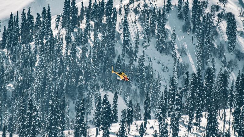 A yellow helicopter flying low over snowy fir forest below a great white mountain face