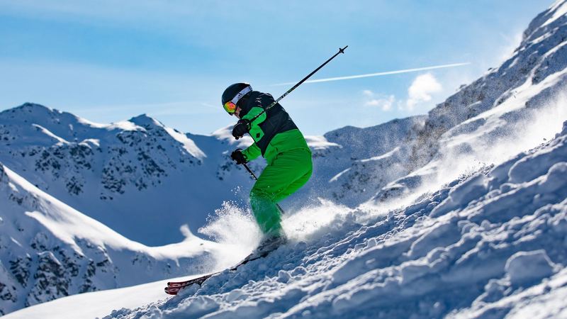 A skier in green carving through powder on a steep slope, snowy peaks against a blue sky