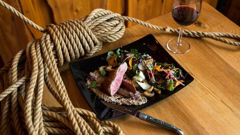 A pink fillet of beef with mushrooms and a crisp salad on a black plate, a glass of red wine on the wooden table