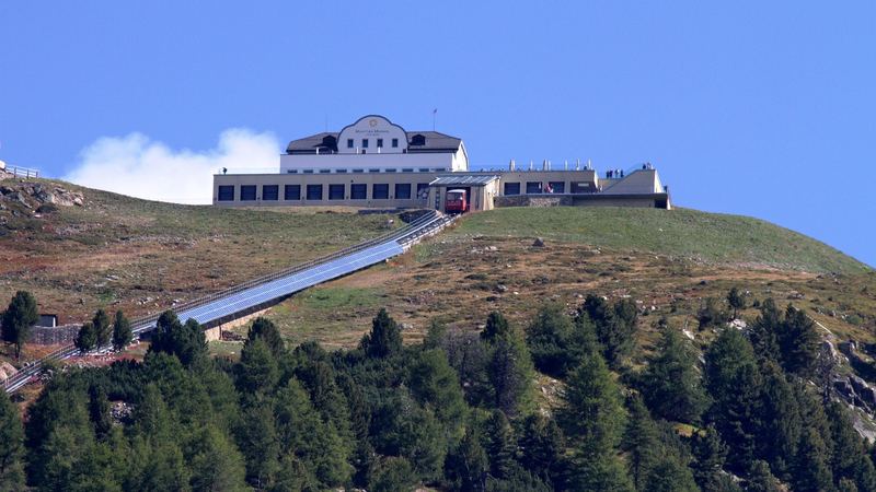 The hotel and terrace of Muottas Muragl on its summit, with the red funicular arriving through the larch forest