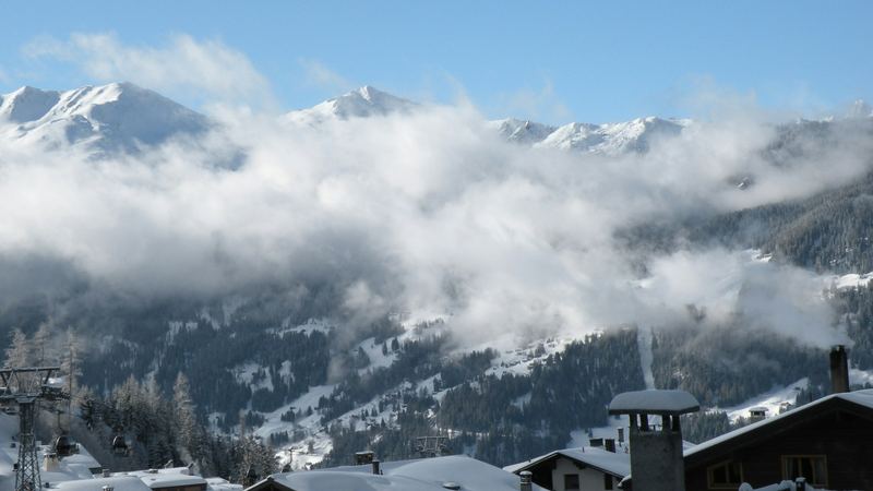 Snow-laden chalet roofs in Verbier, with cloud drifting across the forested mountains beyond