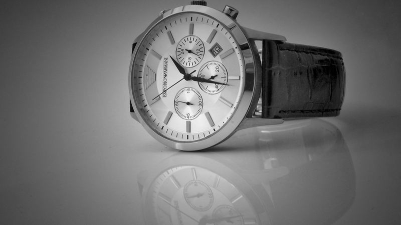 A silver chronograph on a black leather strap, reflected on a pale surface