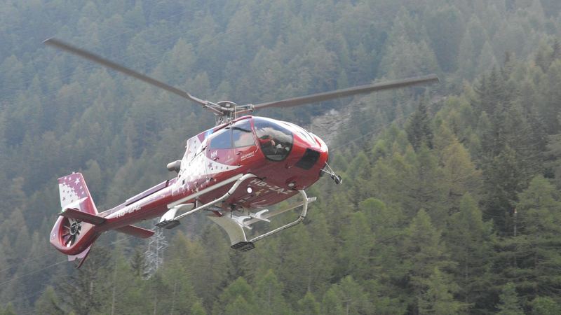 A red Air Zermatt helicopter lifting off in front of a larch forest