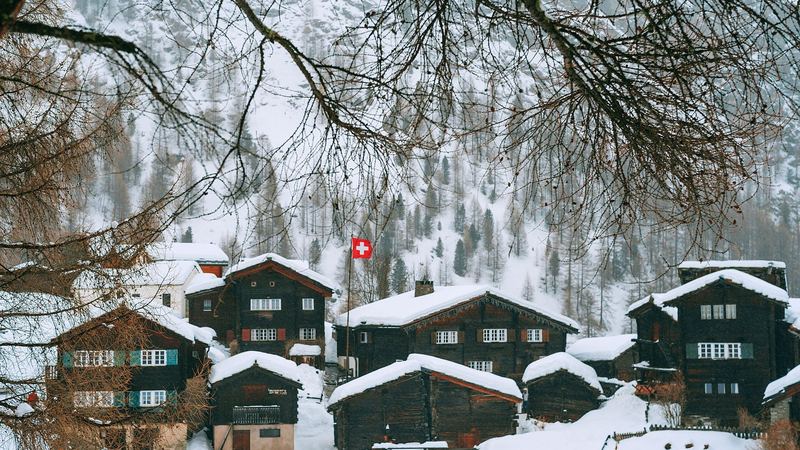 Old timber chalets and barns deep in snow below a forested slope, a Swiss flag among them