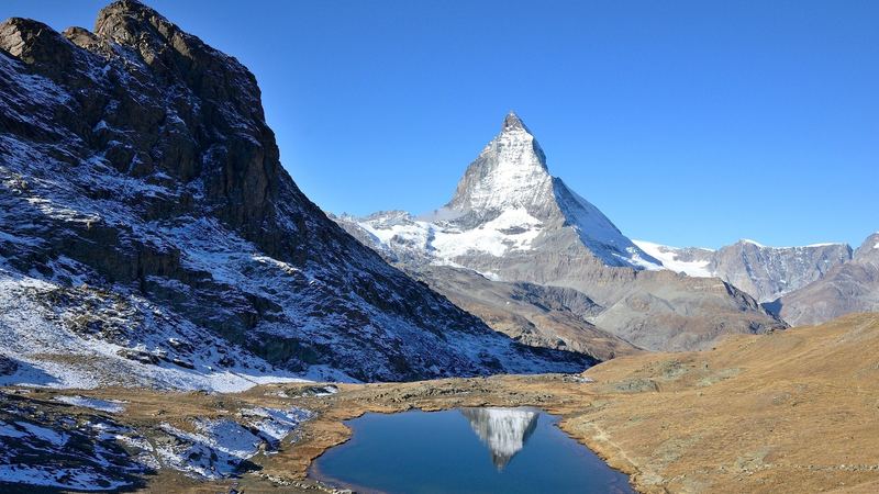 The Matterhorn reflected in the small Riffelsee lake below snow-dusted rocks under a clear blue sky