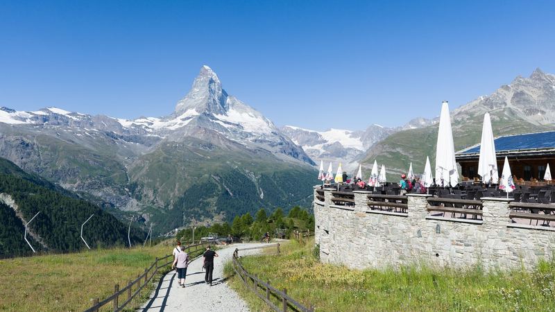 A mountain restaurant terrace with parasols beside a path through the meadows, the Matterhorn beyond